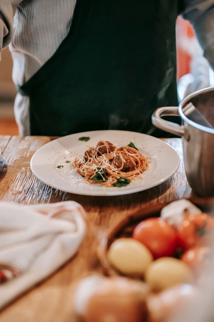 who-we-are Crop unrecognizable man wearing apron while standing near counter and cooking pasta with meatballs and herbs on plate near tomatoes and garlic with lemons in light kitchen near pan